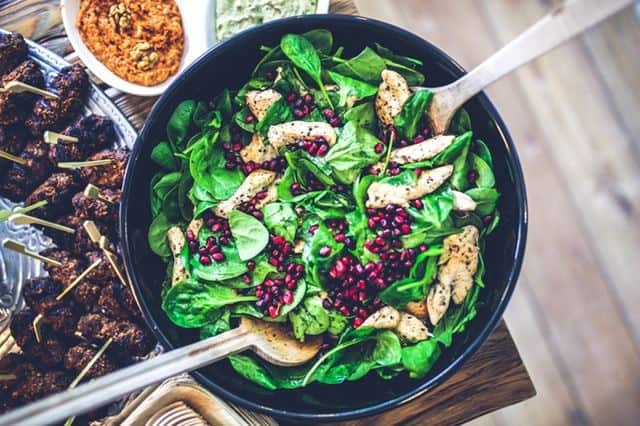 a bowl of salad containing spinach, chicken and pomegranate seeds with salad servers on a table