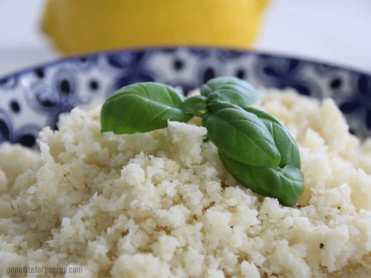 close up of cauliflower rice in a blue bowl with basil leaves on top and a lemon in background