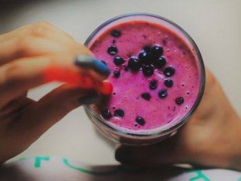 overhead shot of a strawberry smoothie topped with blueberries and a hand holding a red straw about to drink it.