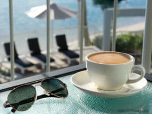 A cup of coffee on table with sunglasses and deckchairs in the background