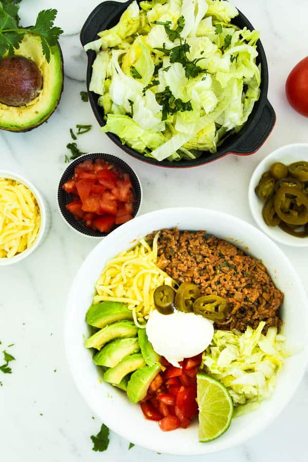 Low catb taco bowls surrounded by bowls of lettuce, chopped tomatoes, cheese and avocado