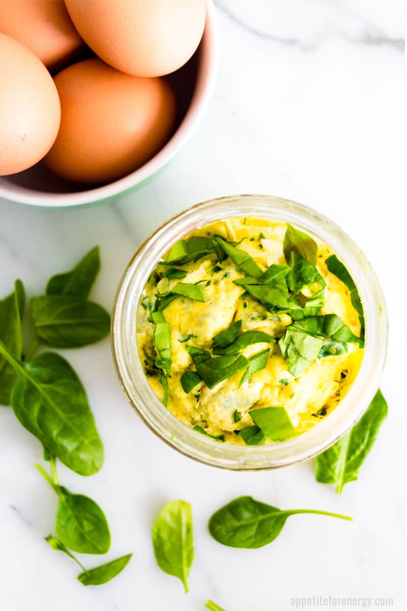 Overhead shot of scrambled eggs in mason jar with basil sprinkled over the top, a bowl of eggs and some basil on the counter