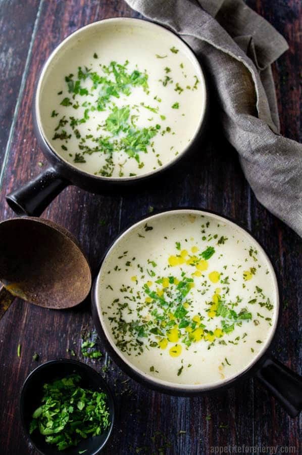 2 bowls of Cauliflower Cheese Soup, sprinkled with herbs and drizzled with olive oil. A wooden ladle, a small bowl of herbs and a beige dish towel are nearby on the table.