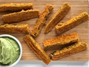 Zucchini Fries scattered on a wooden board with a white bowl containing Spicy Avocado Dipping Sauce
