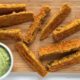 Zucchini Fries scattered on a wooden board with a white bowl containing Spicy Avocado Dipping Sauce