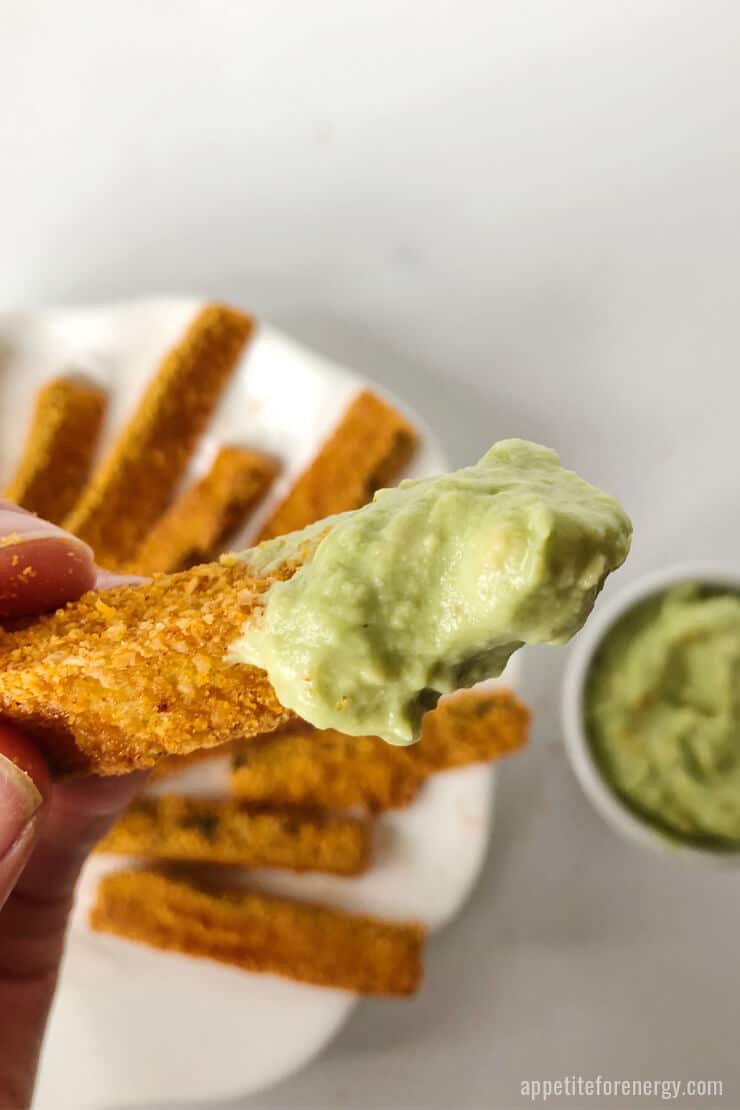 Hand holding a zucchini fry slathered in spicy avocado sauce. A plate of zucchini fries and a bowl of avocado sauce are in the background