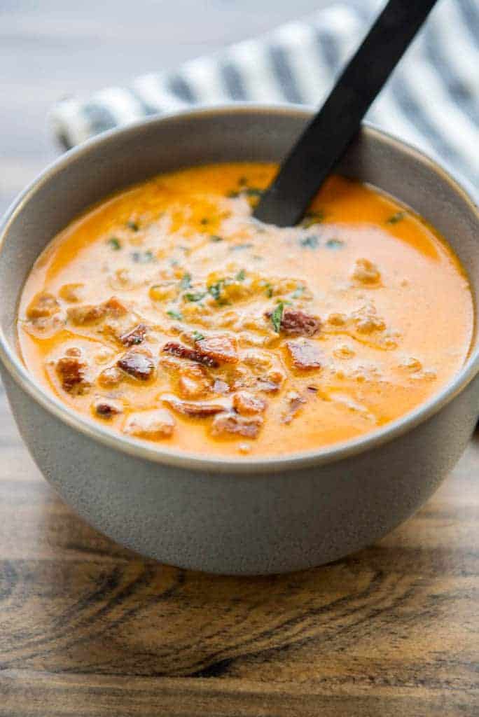 A big bowl of Low Carb Cheeseburger Soup topped with bacon bits and herbs. There is a spoon is in the soup and a dish striped towel in the background