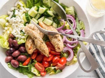 A white bowl with a bed of lettuce, black olives, cherry tomatoes, red onion, cucumber, feta and grilled chicken, Silver tongs