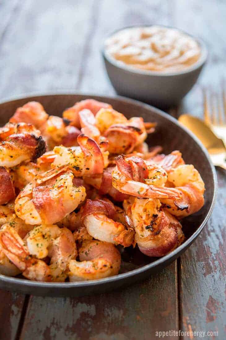 Bacon Wrapped Shrimp in a dark bowl with Remoulade Sauce in a bowl and a gold spoon