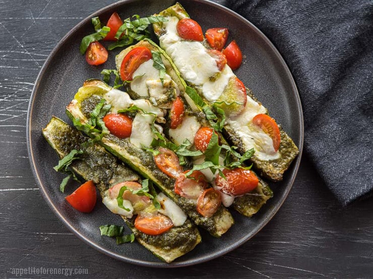 Overhead shot of four Grilled Caprese Zucchini Boats topped with tomatoes, pesto, mozzarella and basil on black plate on a dark table with a grey cloth nearby