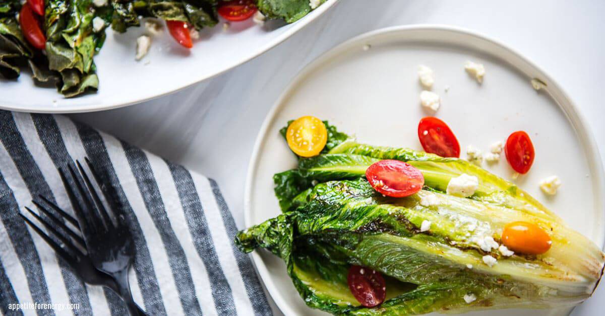 Grilled Romaine on a white plate topped with tomatoes, vinaigrette with a cloth and knife and fork beside