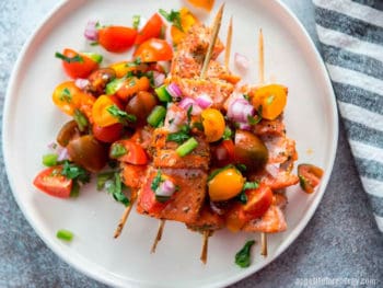 Grilled Salmon Kabobs on a white plate with tomato salsa and fresh herbs scattered on top. Striped cloth nearby