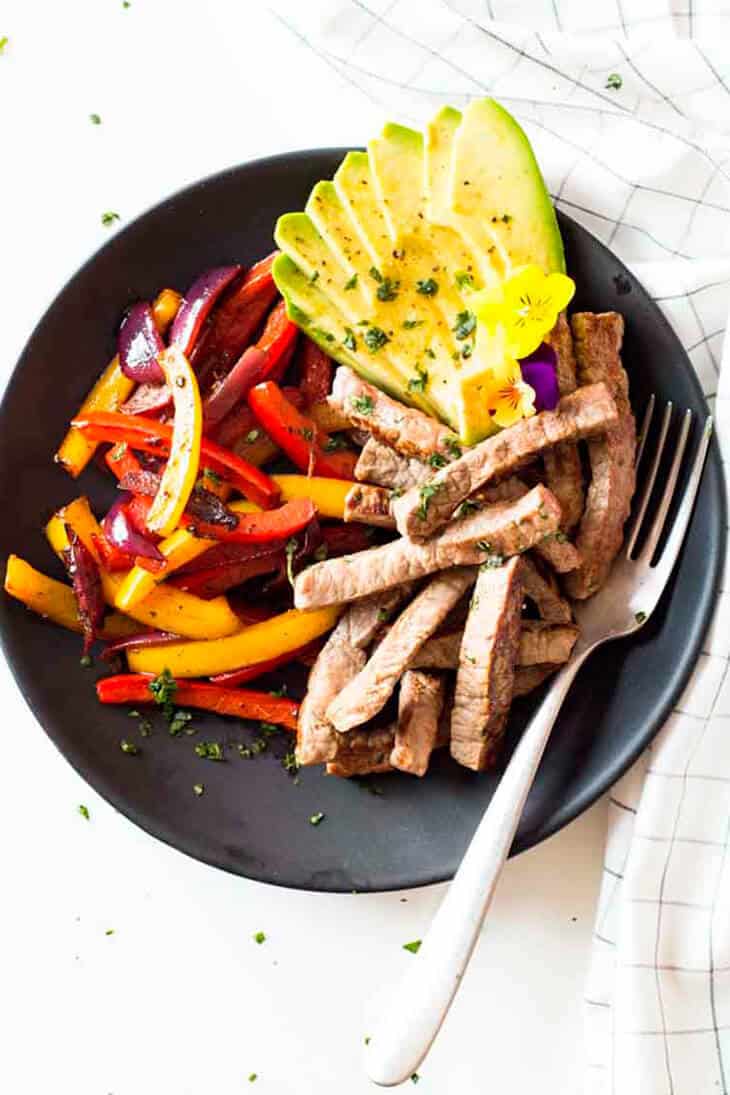 Beef fajitas with red onion, bell peppers and avocado in a black bowl with fork