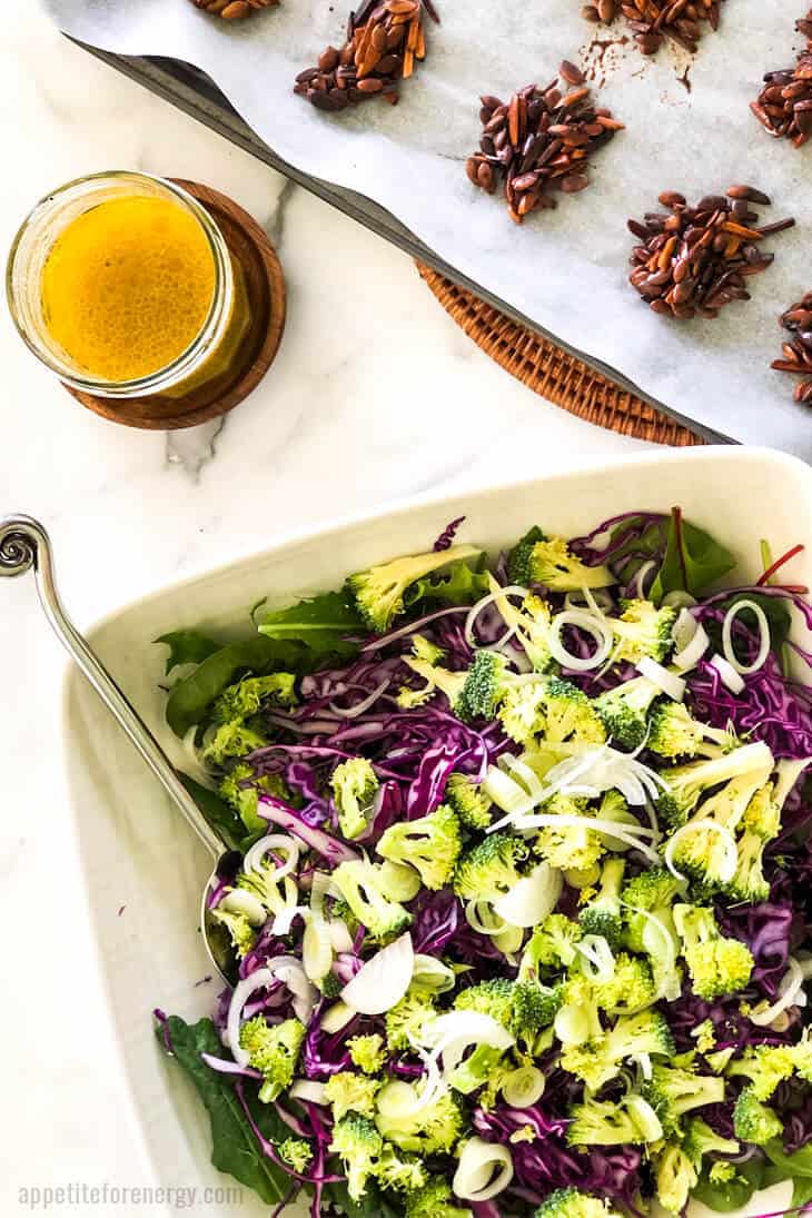 Crunchy Broccoli Slaw in a white serving bowl with nut clusters nearby and dressing in a jar