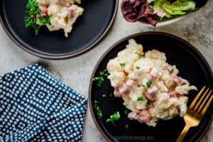 Keto Cauliflower Mac and Cheese with Ham on a black plate with gold fork, lettuce and blue napkin