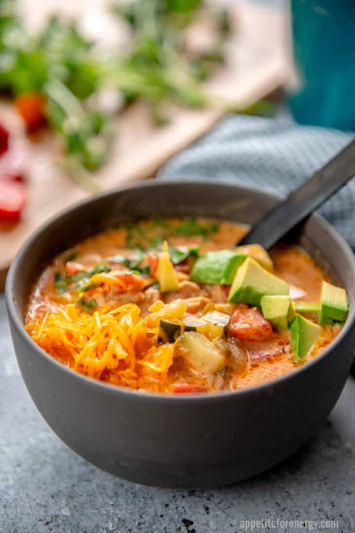 Side view of chicken taco soup in a grey bowl with a ladle. Soup has chunks of avocado, tomato, chicken and shredded cheese.