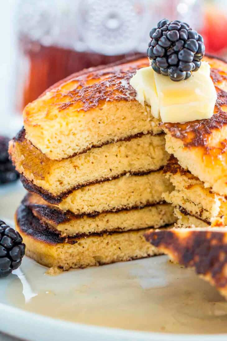 A stack of huge fluffy pancakes with a piece cut out, topped with blackberries and butter