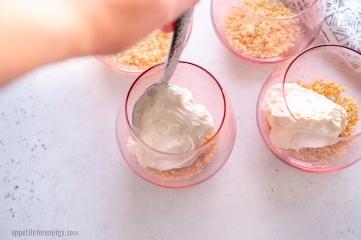 A hand spooning cheesecake mixture into 4 glasses