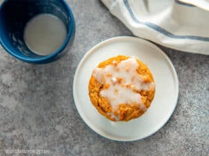 Single low carb cinnamon muffin drizzled with frosting on a white plate and blue bowl of frosting