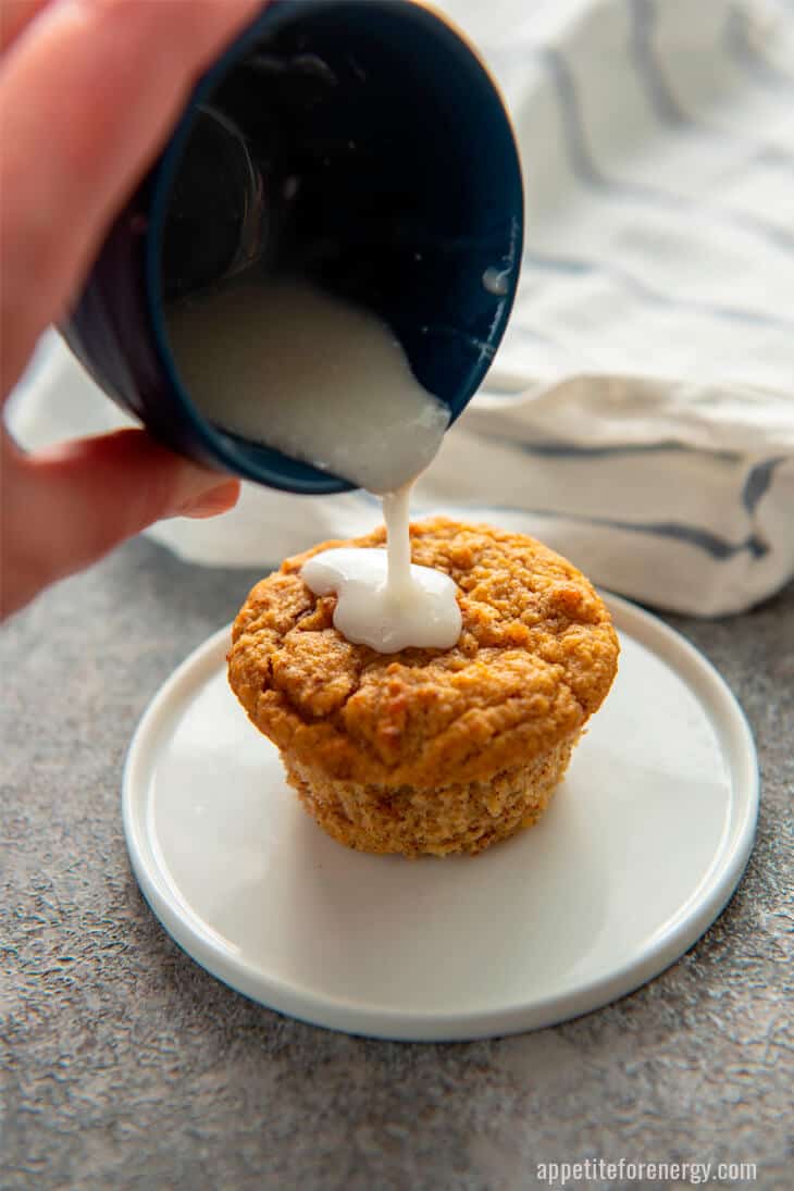 Pouring frosting from blue bowl onto low carb cinnamon muffin on a white plate